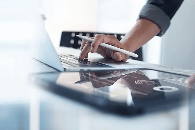 Hand typing on laptop while holding a pen next to a tablet device