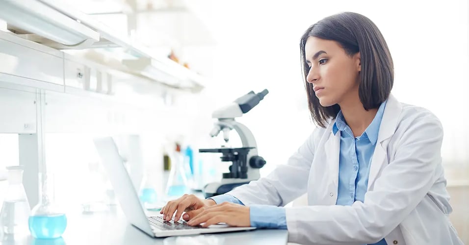 Woman typing on a laptop next to a microscope in a medical laboratory.