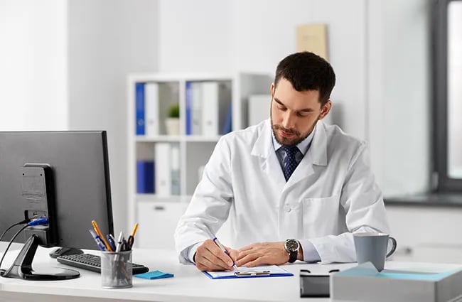 Doctor wearing lab coat writing on clip board at a desk