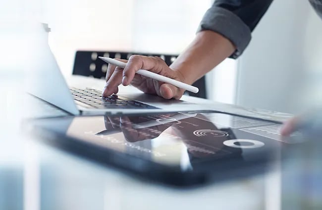 Hand holding pen between fingers typing on laptop and using tablet device