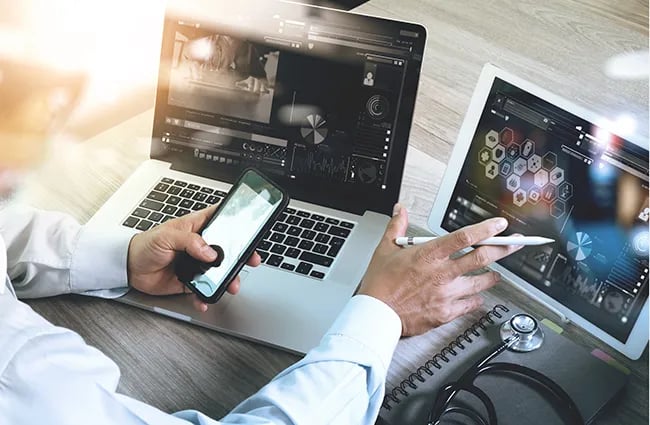 Man using mobile phone tablet and laptop next to stethoscope and spiral notebook