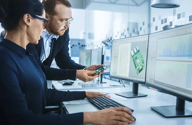 Two engineers reviewing a small computer device in front of two computer monitors