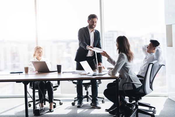 Man standing up handing a stack of papers to a woman in a professional office conference room