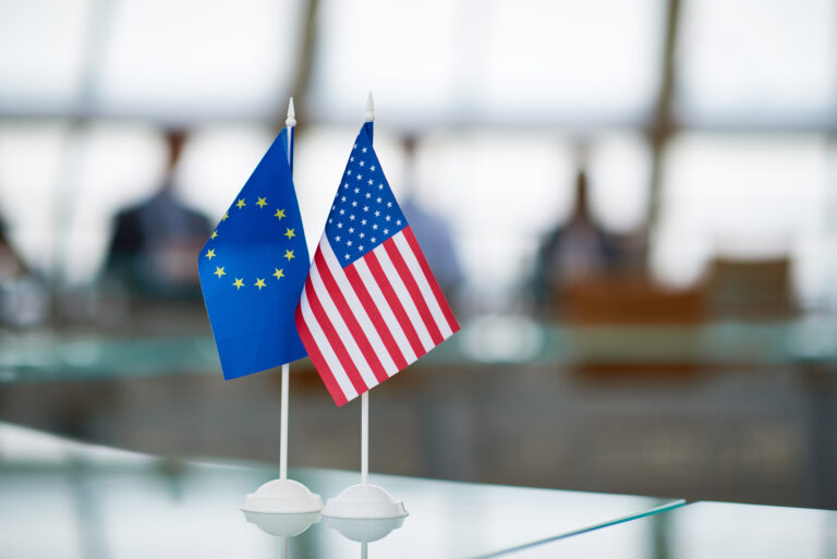 Small flags for the US and EU standing next to each other on a glass desk.