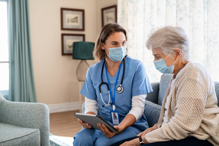 A healthcare professional with face mask on holding a tablet looking at a patient with a face mask on.
