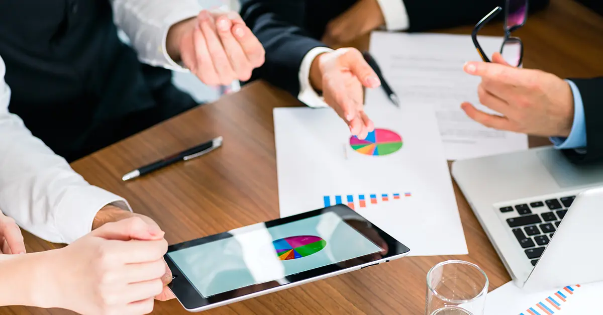 Hands gesturing around a table with papers and tablet devices showing charts and graphs